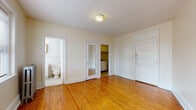 This single-floor unit offers a straightforward and functional layout with timeless design elements throughout. The main living area is bright and open, finished with warm honey-toned hardwood floors that extend through the primary spaces. Neutral, cream-colored walls and a clean white entry door contribute to a light and airy feel. Multiple windows allow natural light to filter in, enhancing the sense of openness.

The kitchenette is efficiently arranged, featuring white cabinetry, a stainless steel sink with countertop workspace, and a full-size refrigerator — providing essential functionality within a compact footprint. A separate doorway from the main room leads directly into this kitchen area.

The bathroom retains classic architectural character, showcasing traditional white subway tile walls, mosaic tile flooring, a pedestal-style vanity, and a cast-iron radiator positioned beneath a bright window with blinds. The vintage radiator heating system is also present in the main room, underscoring the building's period charm.

A mirrored interior door adds a practical touch to the layout, contributing depth and light reflection. The space is well-suited for those seeking a clean, no-frills environment with classic detailing, hardwood flooring, and reliable radiator heat — all contained within a single, accessible level.