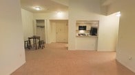 This single-floor residential space offers a functional and comfortable layout designed for everyday living. The open-concept main area seamlessly connects the living and dining zones, providing a generous, flexible space ideal for both relaxing and entertaining. Soft wall-to-wall carpeting in warm neutral tones runs throughout, contributing to a cozy and welcoming ambiance.

The kitchen is accessible via a built-in pass-through window framed by a half-wall, allowing visual connectivity between the kitchen and the main living area while maintaining a sense of separation. The kitchen is equipped with essential appliances, including a refrigerator and microwave, and features light wood cabinetry with under-cabinet lighting that adds warmth to the cooking space.

Adjacent to the main living area is a dedicated dining nook with a compact table and bar-style seating, making it a practical spot for casual meals. A large walk-in closet with a rod and ample hanging space provides generous storage, adding to the unit's overall practicality.

The front entry door opens directly into the main living space, creating an efficient and straightforward floor plan. Ceiling-mounted light fixtures illuminate the interior, and the overall neutral color palette throughout lends the space a clean, move-in-ready aesthetic.