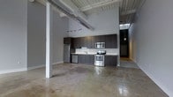 This single-floor space showcases a striking industrial aesthetic defined by soaring ceilings with exposed wooden beams, visible ductwork, and structural white-painted support columns that add to the loft's architectural character. The open-concept layout creates a seamless flow throughout the space, anchored by gleaming polished concrete floors that reflect light and enhance the sense of spaciousness.

The kitchen is thoughtfully designed with a full run of dark espresso cabinetry extending along one wall, offering generous storage both above and below the countertops. Stainless steel appliances — including a refrigerator, electric range, built-in microwave, and dishwasher — lend a modern, functional edge to the space. A single-basin sink with a clean countertop workspace completes the kitchen setup.

Track lighting mounted along the ceiling provides adjustable illumination throughout the open area, while pendant lighting is visible deeper in the space, adding warmth and depth. A corridor visible in the background hints at additional areas beyond the main open floor, further adding to the unit's sense of scale. The overall ambiance is modern and urban, blending raw industrial elements with contemporary finishes for a refined yet edgy living or working environment.