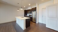 Located at 1252 Osgood in North Andover, Massachusetts, this single-floor residence offers a thoughtfully designed layout with modern finishes throughout. The kitchen serves as a central highlight, featuring dark espresso cabinetry, stainless steel appliances including a refrigerator, range, and built-in microwave, a subway tile backsplash, and a white-topped island with an undermount sink and stylish pendant lighting overhead. Rich, dark hardwood-style flooring flows through the kitchen and living areas, creating a cohesive and contemporary feel. The living area benefits from vaulted ceilings and track lighting, enhancing the sense of openness, while a large window invites natural light. Carpeted bedrooms provide a comfortable, quiet retreat, each with windows offering outdoor views and ample wall space. The primary bathroom is well-appointed with a long dual-sink vanity featuring dark cabinetry and a light countertop, a large mirror, and hardwood-style flooring, with direct access to a spacious walk-in closet complete with built-in shelving. A secondary bathroom includes a combined tub and shower with clean white surroundings, a toilet, and a matching dark vanity. Clean neutral walls and consistent trim work throughout lend the space a polished, move-in-ready appearance.