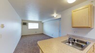 This single-floor residential unit offers a straightforward, functional layout that maximizes its available space through an open-concept design. The living area is finished with wall-to-wall carpeting in a neutral tone, creating a warm and cohesive feel throughout. A wall-mounted air conditioning unit is positioned near a window, ensuring climate comfort year-round, while a floor vent provides additional ventilation. The window allows natural light to filter into the living space, keeping the area bright and inviting.

The kitchen flows seamlessly from the living area and features light wood cabinetry that provides ample storage. A laminate countertop with a stainless steel sink offers a practical workspace, making the kitchen both efficient and easy to maintain. The open pass-through design between the kitchen and living area enhances the sense of space and allows for easy interaction between the two zones.

A white interior door visible from the main living area leads to additional rooms within the unit, suggesting further private spaces beyond the open living and kitchen area. The walls and ceilings are finished in a clean, neutral white, contributing to a simple and tidy ambiance. Overall, the unit presents a clean, move-in ready space ideal for those seeking a practical and comfortable single-floor residence.