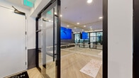 Located at Novel Lakewalk in Sanford, Florida, this contemporary fitness studio is thoughtfully designed to support a full range of workout disciplines within a single, well-organized floor. The space is accessed through striking black-framed glass doors, immediately setting a modern, upscale tone. Light wood flooring runs throughout, complementing the crisp white walls accented with two-tone gray wainscoting.

The studio is divided into distinct functional zones. A dedicated cycling area features a row of stationary spin bikes arranged beneath stylish pendant lighting, creating an energetic atmosphere for group fitness classes. Adjacent to this is a versatile open studio space lined floor-to-ceiling with mirrors, a ballet barre, and a large display screen mounted on a tripod — ideal for yoga, barre, stretching, or virtual instructor-led sessions. The mirrored walls amplify the sense of space and light throughout.

Glass partition walls and railings with warm wood-capped handrails provide visual connectivity between zones while maintaining separation. A rack of stability balls and additional fitness accessories rounds out the available equipment. Recessed and pendant lighting ensures the entire studio is well-lit. The overall design balances form and function, creating an inviting, professional-grade fitness environment.