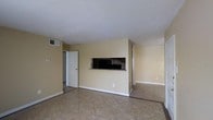 This single-story space presents a clean, functional layout centered around an open living area. The room is finished with neutral beige walls and large-format tile flooring in a warm tan tone, contributing to a bright and easy-to-maintain environment. A standout architectural detail is the built-in wall niche, framed with stacked stone tile accenting and a dark interior, providing a designated space for a television or display piece. This feature adds a touch of structured design to an otherwise simple interior. Overhead, a standard white ceiling hosts a centrally placed HVAC vent, ensuring climate control throughout the space. White baseboards run along the perimeter, offering a clean contrast against the tiled floors. A white door leads to an adjacent room or closet, while an open passageway to the right connects to additional areas of the unit. The overall ambiance is minimal and neutral, providing a blank canvas that lends itself well to a variety of furniture arrangements and interior design preferences. The space feels open and uncluttered, making it adaptable for everyday living.