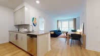 This single-floor residence is designed with a clean, contemporary aesthetic that emphasizes an open, airy atmosphere. The heart of the unit is the open-concept living area, where the kitchen, dining, and living spaces flow seamlessly together. The kitchen is outfitted with white shaker-style cabinetry extending to the ceiling, stainless steel appliances including a French door refrigerator, microwave, and range, and a peninsula with a taupe quartz countertop that provides both additional prep space and casual seating. A stainless steel dishwasher is integrated beneath the peninsula.

The living area benefits from generous natural light provided by large floor-to-ceiling windows, with views to the outdoors. Light oak hardwood flooring runs consistently throughout the main living spaces, enhancing the sense of openness. A dedicated dining area sits adjacent to the living room, which features a wall-mounted television and ample space for seating.

The primary bedroom is calm and well-proportioned, with a window providing natural light and room for a full furniture arrangement. A secondary bedroom offers built-in wardrobe storage and versatile use as a child's room or additional sleeping space. Two bathrooms serve the unit: one featuring a glass-enclosed shower and tub combination with white subway tile, and a second with a vanity and full mirror. Both bathrooms maintain the home's crisp, minimalist palette.