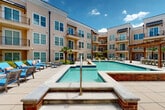 Located at 4301 Tulane Ave in New Orleans, Louisiana, this multi-story residential complex is designed around a striking central courtyard that serves as the heart of the community. The building's exterior features a warm, neutral lap-siding façade with metal accent rooflines, large windows, and private wrought-iron balconies that overlook the shared outdoor amenity space below.

The centerpiece of the courtyard is a generously sized resort-style swimming pool with clear blue water and classic blue tile detailing, complemented by an adjacent raised spa with brick surround and stainless steel entry rails. The pool deck is finished in large-format stone pavers and furnished with lounge chairs and seating areas ideal for relaxation and socializing.

Lush tropical landscaping, including mature palm trees and manicured garden beds, adds a vibrant, subtropical character fitting of New Orleans' climate. A pergola structure at one end of the courtyard provides a shaded gathering area, while additional patio seating is distributed throughout the space for resident use.

Separate paved entry courtyards with ground-floor sliding glass doors and upper-level balconies create a layered, accessible layout throughout the building. The architecture strikes a balance between contemporary design and Southern residential character, making this a well-appointed and visually cohesive property in the heart of New Orleans.