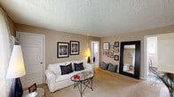 Located at Patterson Village in Dayton, Ohio, this one-bedroom, one-bathroom unit offers a practical and well-organized floor plan across a single level. The living room is the heart of the space, featuring warm neutral-toned walls, plush carpeting, and a textured ceiling. Natural light filters through sheer curtains, complemented by a wall-mounted air conditioning unit. The room offers ample space for seating and storage, with built-in shelving along one wall providing display and organizational options.

The kitchen is efficiently designed with white cabinetry extending to the ceiling for maximum storage, light granite-style countertops, wood-look flooring, and full-size appliances including a refrigerator and microwave.

The bedroom is a comfortable retreat, featuring carpeted floors, a window with sheer curtains that invite natural light, and an impressive set of overhead wall-mounted white cabinets that maximize vertical storage. Neutral gray walls create a calm atmosphere.

The bathroom includes a full bathtub and shower combination, accented by retro warm yellow and terracotta wall tiles, with a window providing natural ventilation. A connecting hallway ties all rooms together, creating a cohesive and functional living environment.