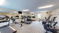 Located at 1151 Gregory Mills Dr in Smyrna, Tennessee, this fitness center offers a well-organized, single-floor exercise space designed to support a variety of workout routines. The room features durable patterned carpet flooring throughout, neutral-toned walls, and a textured ceiling fitted with bright fluorescent lighting that ensures the space is well-lit at all times. Multiple windows along the walls invite natural light and provide views of the surrounding greenery, contributing to an open and energizing atmosphere. A large wall mirror spans one side of the room, visually expanding the space and allowing users to monitor their form during workouts. The facility is equipped with multiple treadmills, including Endurance-brand models, as well as elliptical machines, an air resistance bike, a cable strength machine, and a weight bench, offering a comprehensive range of cardio and resistance training options. A wall-mounted flat-screen television is positioned for easy viewing during exercise sessions. Additional amenities include a wall-mounted equipment station and a waste bin for convenience. The layout is thoughtfully arranged to maximize floor space and allow comfortable movement between equipment. With its practical design, ample equipment, and clean, functional environment, this fitness center provides everything needed for an effective daily workout.