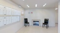 This single-floor lobby serves as a well-appointed communal entrance space, thoughtfully designed to balance functionality with comfort. The space is finished with light-toned floor tiles that lend a clean, bright feel throughout. Recessed and flush-mounted fluorescent lighting illuminates the area evenly, ensuring a well-lit environment at all times.

A standout feature of the lobby is the built-in fireplace, framed in a neutral surround, which anchors one wall and adds a warm, welcoming ambiance to the space. Flanking the fireplace are two black leather accent chairs, providing a simple and practical seating arrangement for residents and guests.

Along the adjacent wall, a comprehensive bank of individual resident mailboxes is neatly mounted, offering organized mail and parcel access for building occupants. Beside the mailboxes, a cork bulletin board serves as a central information hub for community notices and announcements. A wall-mounted intercom or access control panel is also present, supporting building security and visitor management.

The overall design of the lobby is clean and modern, with neutral wall tones and minimal ornamentation that create a professional and functional atmosphere. This entrance space effectively combines essential building services with a comfortable setting for residents.