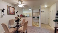 This single-floor residence offers a thoughtfully designed open-concept layout that promotes a comfortable and functional living experience. The dining area is anchored by a round glass-top table with cushioned chairs, set beneath a ceiling fan with integrated lighting that enhances air circulation and ambiance. Warm, neutral-toned carpeting runs throughout the main living and dining spaces, creating a cozy and inviting atmosphere.

The kitchen is positioned adjacent to the dining area and is defined by a half-wall pass-through counter, providing both visual connection and practical separation. Light wood cabinetry lines the kitchen walls, offering ample storage, while a built-in microwave and standard appliances complete the space. The kitchen features a distinctive checkered tile floor in soft yellow and cream tones, adding a classic, charming character.

Beyond the kitchen, a dedicated utility nook with additional cabinetry and counter space provides extra functionality, ideal for use as a workspace or secondary storage area. A set of bifold closet doors nearby adds further organizational convenience. The overall palette of soft whites, warm beiges, and light wood tones gives the entire space a bright, clean, and welcoming aesthetic throughout.