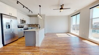 This contemporary single-floor unit offers a thoughtfully designed open-concept layout that seamlessly connects the kitchen and living area. Rich hardwood flooring extends throughout the main living space, complemented by neutral-toned walls that enhance the bright, airy atmosphere. The kitchen is a standout feature, showcasing two-tone cabinetry with crisp white upper cabinets and slate-blue lower cabinets, light stone countertops, a subway tile backsplash, stainless steel appliances, and a central island with a built-in sink — ideal for both cooking and entertaining. A ceiling fan and track lighting add functional style overhead. Large floor-to-ceiling windows in the living area flood the space with natural light and offer open outdoor views. The bedroom continues the bright aesthetic with plush carpeting, a ceiling fan, and an oversized window framing skyline views. The primary bathroom is well-appointed with large-format tile flooring, a white double vanity with matte black hardware, a glass-enclosed shower, and direct access to a spacious walk-in closet with built-in shelving and hanging rods. A secondary bathroom provides clean, modern finishes with a single vanity and tile flooring. Throughout, matte black accents and thoughtful detailing lend a cohesive, contemporary character to the entire space.