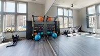 Located at 9235 Senator Royall Dr. in Charlotte, NC, this single-floor fitness center is thoughtfully designed to support a comprehensive workout experience. The space is defined by soaring, floor-to-ceiling windows that line multiple walls, flooding the interior with natural light and offering open views of the surrounding community. The tall ceilings and expansive glazing lend the gym a bright, airy atmosphere that feels spacious and welcoming.

The flooring throughout is a durable wood-look material in a neutral grey tone, complementing the modern, neutral-toned walls. A ceiling fan provides additional air circulation during workouts. The facility is equipped with Matrix-branded cardio machines, including stationary bikes and a rowing machine, as well as a dedicated cycling area. A large wall-mounted mirror amplifies the sense of space and supports strength and form training.

Functional fitness equipment is well-represented, with a multi-purpose rack system holding an assortment of kettlebells, medicine balls, and stability balls, alongside free weights and exercise mats. A large flat-screen display is mounted near the entrance, adding a modern multimedia element to the space. Access is secured via a contemporary paneled door with a keypad entry system, ensuring a private and controlled environment for residents.
