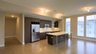 This single-floor space showcases a contemporary open-concept layout centered around a well-appointed kitchen. Dark espresso-toned cabinetry lines the walls, extending from floor to ceiling to maximize storage, while stainless steel appliances — including a French door refrigerator with an ice dispenser, a built-in microwave, and a range — provide a sleek, professional aesthetic. Open shelving above the countertops adds a modern touch and practical display space.

The kitchen island serves as a focal point, featuring a clean white countertop that contrasts beautifully with the dark cabinetry below. An extended overhang supported by metal legs creates a generous breakfast bar, ideal for casual dining or entertaining. Pendant lighting hangs above the island, complementing the track lighting installed overhead to ensure the workspace is well-illuminated.

Light hardwood flooring flows throughout the open space, adding warmth and continuity to the modern design palette of neutral beige walls. A tall vertical window adjacent to the kitchen allows natural light to filter in, enhancing the bright and airy atmosphere of the space.