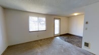This single-floor residence offers a straightforward and functional layout designed for comfortable everyday living. The main living area is a welcoming, open space that benefits from abundant natural light streaming in through a large front-facing window fitted with vertical blinds, offering views of the surrounding outdoor greenery. A secondary door with a glass panel provides direct access to the exterior, further enhancing the bright and airy feel of the room. The living area features a combination of plush carpeting and durable tile flooring, creating a practical transition zone near the entryway. Neutral, light-colored walls complement the space throughout, lending it a clean and versatile aesthetic that adapts easily to various décor styles. The textured popcorn ceiling adds a classic residential character, while flush-mounted ceiling lighting ensures the space remains well-lit. Dark baseboards provide a subtle contrast to the pale walls, adding a touch of definition to the room's boundaries. Overall, the space presents a functional and low-maintenance environment, ideal for those seeking a simple, comfortable, and well-lit living arrangement on a single level.