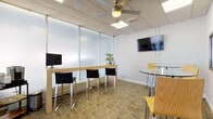 This single-floor office space is thoughtfully designed to support a modern, collaborative work environment. The layout is open yet organized, combining distinct functional zones within a cohesive interior. A drop ceiling with recessed panel lighting and a ceiling fan runs throughout, providing consistent illumination and airflow across the space.

The main area features patterned commercial carpeting and crisp white walls accented by vibrant cityscape artwork. High-top glass bistro tables paired with sleek, yellow bentwood barstools create informal gathering spots ideal for quick meetings or casual collaboration. A wall-mounted flat-screen display serves as a focal point for presentations or shared content.

A dedicated workstation area includes a long natural wood-toned counter fitted with monitor mounts and black upholstered barstools, positioned against frosted glass partitions that diffuse light while maintaining privacy. Adjacent to this, a small refreshment station with a coffee maker and shelving adds practical convenience.

Glass-paneled interior walls and a full-glass entry door connect the space to neighboring office suites, enhancing visual openness while clearly delineating boundaries. Potted plants are placed throughout, adding greenery and a welcoming atmosphere. The overall design balances functionality and style, creating a professional yet approachable workspace.