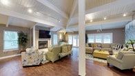 This single-story space showcases an impressive open-concept layout that seamlessly connects the dining and living areas beneath dramatic vaulted ceilings with white-painted shiplap and exposed structural beams. A row of clerestory windows along the roofline floods the interior with abundant natural light, complementing the warm-toned hardwood flooring that runs throughout.

The dining area features a rustic wood dining table surrounded by upholstered chairs with nailhead trim detailing, anchored by a bold yellow patterned area rug. A large floral painting adds a decorative focal point to the space.

The expansive living area is divided into two distinct seating zones. The first centers around a shiplap-clad fireplace with a wall-mounted flat-screen television above, flanked by comfortable sofas and accent chairs in soft greens, blues, and floral patterns. The second zone features a large sectional sofa with coordinating yellow ottoman, positioned to take advantage of the multiple sets of French doors that open to the outdoors.

White structural columns define zones without interrupting the open flow. Track lighting and wall sconces provide warm ambient illumination throughout. The overall aesthetic is coastal-inspired, combining neutral tones with cheerful yellow and turquoise accents for a relaxed yet polished ambiance.