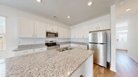 Located at 2108 Silly Soil Ct in Leland, NC, this two-story home offers a thoughtfully designed layout spread across two floors. The main level features an open-concept kitchen and living area anchored by a stunning kitchen with granite countertops, a central island, crisp white shaker cabinets, and stainless steel appliances. Light wood-look flooring runs throughout, complementing the clean, neutral palette. The living room is bright and airy, highlighted by a ceiling fan and sliding glass doors that open to an outdoor view, filling the space with natural light. A garage with an automatic door provides convenient covered parking and additional storage. Upstairs, the primary bedroom is generously sized and bathed in natural light through multiple windows, with a ceiling fan adding comfort. Two additional bedrooms feature large windows with outdoor views and the same warm wood-look flooring. A well-appointed full bathroom with granite vanity, tub-shower combo, and white cabinetry serves the upper level. A dedicated laundry room with wire shelving and washer/dryer hookups adds everyday convenience. The home also includes a hallway closet for extra storage, making it as functional as it is attractive.