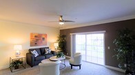 This single-floor residence offers a thoughtfully designed living space that balances comfort and style. The living area serves as the heart of the home, featuring plush carpeting underfoot and a ceiling fan with integrated lighting that enhances the room's airy feel. A rich, dark brown accent wall adds depth and visual interest, complementing the neutral tones found throughout the space.

Large sliding glass doors flood the living area with abundant natural light while providing direct access to an outdoor balcony, where views of the surrounding greenery can be enjoyed. The open layout accommodates a generous seating arrangement, including a sofa, accent chairs, and a central coffee table, making the space ideal for both relaxing and entertaining.

Warm, ambient lighting from table lamps contributes to an inviting atmosphere, while contemporary wall art adds a pop of color and personality. Lush indoor plants placed strategically near the balcony doors further connect the interior to the outdoors, enhancing the overall sense of freshness and vitality. Clean white ceilings and trim provide a crisp contrast to the warmer tones of the walls, creating a well-balanced and welcoming environment throughout this single-level home.