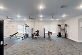 Located at 1301 Rudel Drive in Tomball, Texas, this fitness center offers a comprehensive workout facility spread across two interconnected exercise rooms on a single floor. The space is thoughtfully designed with clean white walls, neutral gray patterned carpet flooring, and recessed lighting complemented by ceiling fans that maintain a comfortable, well-ventilated environment throughout.

The first exercise room is dedicated to cardio training, featuring multiple treadmills and an elliptical machine positioned near bright, white-shuttered windows that allow ample natural light to fill the space. The second, larger room is outfitted with a variety of strength-training equipment, including multi-station cable machines, seated weight machines, and free-weight benches, providing a well-rounded resistance training experience.

A transitional open area connects the two rooms and serves as a functional flex space, housing a wall-mounted medicine ball rack with a colorful selection of weighted balls for dynamic training, as well as a beverage vending machine for post-workout refreshment. Vibrant fitness-themed artwork is displayed throughout the rooms, adding an energetic and motivating aesthetic. The facility's bright, airy atmosphere and thoughtful equipment layout make it a practical and inviting space for a full-body fitness routine.