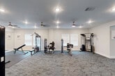 Located at 1301 Rudel Drive in Tomball, Texas, this fitness facility offers a thoughtfully designed workout environment across two dedicated exercise rooms on a single floor. The space is finished with neutral gray carpet tiles throughout, crisp white walls, and clean white baseboards, creating a bright and motivating atmosphere.

The first exercise room is dedicated to strength training and houses a range of cable and resistance machines, including multi-use weight stations and isolated muscle machines. Ceiling fans with integrated lighting complement the recessed lighting overhead, while white-framed windows allow natural light to filter in. Colorful wall art adds an energetic touch to the space.

The second exercise room focuses on cardio equipment, featuring multiple treadmills and an elliptical machine arranged along windowed walls to allow for natural light during workouts. A ceiling fan keeps air circulating throughout.

A connecting corridor area provides open floor space for functional training, with a wall-mounted medicine ball rack offering a variety of weighted balls. A beverage vending machine is conveniently located in this transitional space. Additional storage shelving is also available nearby. Together, the two rooms form a comprehensive, well-organized fitness center suitable for a full range of workout routines.