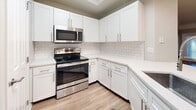 This 980-square-foot, single-floor apartment at 10135 Gate Parkway N. in Jacksonville, Florida offers a well-designed 2-bedroom, 2-bathroom layout. The kitchen is a standout feature, showcasing white shaker cabinetry, subway tile backsplash, light quartz countertops, and a full suite of stainless steel appliances including a range, over-the-range microwave, and refrigerator. A peninsula with a built-in sink provides additional prep space and casual dining potential. Wood-look vinyl flooring flows consistently throughout the entire unit, adding warmth and cohesion. The open living area features crown molding and a ceiling fan, while large windows in the bedrooms invite natural light. The primary bedroom, at 150 square feet, includes a ceiling fan and direct access to a walk-in closet and a private en-suite bathroom with a tiled walk-in shower. The second bathroom features a full tub-shower combo and a white vanity, serving both guests and the second bedroom. Two hallways and two closets offer practical storage solutions throughout. The unit presents a clean, neutral color palette with modern finishes, creating a bright and functional living environment.