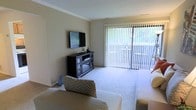 This single-floor space centers around a well-appointed living room designed for both comfort and functionality. The room is finished with plush wall-to-wall carpeting in a neutral tone, contributing to a warm and inviting atmosphere. Soft, beige walls complement the overall light color palette, while crown molding along the ceiling adds a subtle architectural detail that lends a polished finish to the space.

A wall-mounted flat-screen television serves as a focal point, positioned above a dark wood media console that provides both display space and storage for media components. Seating includes a cushioned sofa accented with a patterned throw pillow, offering a relaxed and casual arrangement.

Natural light is a standout feature of this living room, made possible by a large sliding glass door fitted with vertical blinds. The door opens to what appears to be an outdoor balcony or patio, offering views of surrounding greenery and allowing ample daylight to fill the room. The combination of natural light, neutral finishes, and functional furnishings creates a balanced and livable environment suited to everyday use.