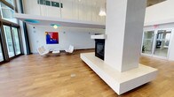 This contemporary space makes an immediate impression with its dramatic double-height ceilings and expansive open-plan layout. The central architectural feature is a freestanding, floor-to-ceiling white fireplace column with a modern built-in firebox, serving as both a functional focal point and a bold design statement that elegantly divides the floor plan. Warm hardwood flooring runs throughout, providing a natural contrast to the crisp white walls and clean-lined finishes. The seating area is furnished with sleek, minimalist lounge chairs arranged around a low coffee table, complemented by a vibrant large-scale abstract painting in bold reds and blues that adds a dynamic splash of color to the otherwise neutral palette. Above, an upper-level mezzanine with glass railings overlooks the main floor, contributing to the sense of openness and vertical scale. Pendant lighting with cascading elements descends from the ceiling, while teal accent light fixtures add subtle pops of color. Recessed lighting and generous glazing ensure the space remains bright and airy throughout. The overall ambiance is one of sophisticated, gallery-like modernism — refined, open, and visually engaging.