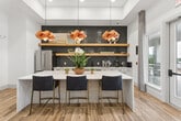 Located at 4770 Military Drive West in San Antonio, TX, this single-floor space offers a thoughtfully designed open-concept layout that seamlessly connects the living room, dining area, and kitchen. The living room is a standout feature, anchored by a generous sectional sofa, warm leather accent chairs, and a built-in shelving unit with a large wall-mounted TV. Tall windows dressed in layered sheer and linen curtains flood the space with natural light while maintaining an air of elegance. The kitchen is a design highlight, boasting a long quartz-topped island with seating for multiple guests, striking navy blue decorative tile backsplash, open wood floating shelves, stainless steel appliances, and sculptural copper pendant lighting that adds warmth and character. The dining area features a round marble-top table paired with sleek navy blue chairs, set against more floor-to-ceiling draped windows and a vibrant piece of wall art. Throughout, light wood-look flooring ties each zone together cohesively. The neutral palette is punctuated by curated artwork and greenery, creating a polished yet inviting atmosphere suited for both relaxed everyday use and entertaining.