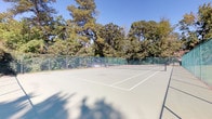 This outdoor tennis facility offers a well-maintained hard court surface with crisp white line markings and a standard net, designed to meet the needs of recreational and competitive players alike. The court is fully enclosed by green chain-link fencing, providing both safety and a visually cohesive aesthetic that blends naturally with the surrounding landscape.

The setting is defined by its lush, mature tree canopy that frames the court on multiple sides, offering partial shade along the perimeter and a tranquil, park-like atmosphere. The open sky above ensures ample natural light across the playing surface throughout the day.

Adjacent to the court, a grassy lawn area features a covered pavilion structure with a distinctive multi-colored roof, along with outdoor seating, making it a practical gathering spot for spectators or players between matches. Additional structures, including what appears to be a brick building, are visible in the background, suggesting this court is part of a broader recreational or community complex.

The combination of a well-kept playing surface, secure fencing, mature greenery, and nearby amenities makes this an inviting and fully functional outdoor sports space suited for regular use and organized play.