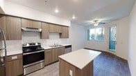 This single-floor residence offers a clean, contemporary design with an open-concept layout that seamlessly connects the kitchen and living areas. The kitchen is a standout feature, equipped with stainless steel appliances including a refrigerator, electric range, and dishwasher. Warm-toned flat-front cabinetry extends from floor to ceiling, providing generous storage, while light quartz countertops offer ample workspace. A central island with a matching quartz surface adds additional prep space and serves as a natural divider between the kitchen and living area. Light wood-look flooring runs throughout the kitchen, transitioning to a darker wood-look finish in the living and sleeping areas. The living space benefits from natural light through windows dressed with horizontal blinds, and a ceiling fan adds comfort overhead. A sliding glass door provides access to an outdoor area. The bedroom is a bright, well-proportioned room with a window and ceiling fan. The bathroom features wood-look tile flooring, a full-size soaking tub with a tiled surround, grab bars for added accessibility, and a toilet. A dedicated laundry area with hookups is also present within the unit. The overall aesthetic is modern and move-in ready.
