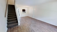 This two-story space offers a well-organized layout anchored by an open main living area on the ground floor. Light-toned hardwood-style flooring runs throughout, lending the space a clean, airy aesthetic that pairs naturally with the bright white walls and white baseboards. A carpeted staircase with a white banister rises along one side of the room, providing a clear transition to the upper level while adding a subtle contrast in texture.

The open living area flows seamlessly toward a dining and kitchen space at the rear of the floor. A chandelier with a classic design hangs overhead near the dining area, serving as a decorative focal point. The kitchen is partially visible through a pass-through opening, featuring dark cabinetry and a raised counter or bar-height surface, offering both a functional workspace and an informal gathering point.

The white textured ceiling and neutral wall tones throughout the main floor contribute to a bright, cohesive atmosphere. The open-plan arrangement of the living, dining, and kitchen areas promotes an efficient use of space and allows for flexible furniture placement, making the ground floor feel connected and welcoming.
