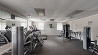 This fitness center offers a thorough and well-organized workout environment designed to support a wide range of exercise routines. The space is laid out on a single floor and features a drop-panel ceiling with integrated lighting and ventilation, complemented by multiple windows fitted with horizontal blinds that allow ample natural light to fill the room. Crown molding adds a subtle architectural detail along the upper walls, lending a clean, finished look to the interior.

The flooring throughout is a durable, dark-toned carpet well-suited for gym use, providing cushioning and stability underfoot. A large wall mirror on one side of the room visually expands the space and aids users in monitoring their form during exercise.

Cardio equipment is well represented, with multiple treadmills, an elliptical trainer, and a recumbent stationary bike positioned throughout the room. Strength training options include a cable machine, a multi-functional weight station, and several selectorized resistance machines targeting various muscle groups. The equipment is arranged to allow clear traffic flow and access throughout the space.

Entry is provided through a set of white French doors, contributing to the room's clean, neutral aesthetic. The overall ambiance is functional and inviting, with a bright, open feel supported by the light-colored walls and consistent natural illumination from the surrounding windows.