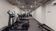 This fitness center offers a well-organized and fully equipped space designed to support a complete workout routine. The open layout accommodates a generous selection of SportsArt cardio machines, including a row of treadmills and ellipticals lined along one wall, complemented by a recumbent bike and additional elliptical trainers for varied low-impact training options. A dedicated free weights area features a multi-tiered dumbbell rack stocked with a full range of weights, paired with an adjustable workout bench ideal for strength training exercises. A large floor-to-ceiling mirror spans one wall, enhancing the sense of space and allowing users to monitor their form during workouts. The flooring throughout is durable rubber gym matting, providing cushioning and stability underfoot. Overhead, multiple ceiling fans circulate air throughout the room, while recessed lighting and exposed ceiling infrastructure lend the space an industrial yet functional aesthetic. A wall-mounted air conditioning unit ensures a comfortable environment year-round. The fitness center is accessed through glass-paneled doors, offering visibility into the space and connecting it to the broader building amenities. Clean white walls and a neutral color palette create a focused, distraction-free atmosphere well-suited for exercise.
