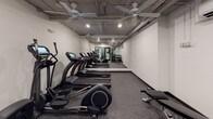 This fitness center offers a well-organized and fully equipped space designed to support a complete workout routine. The open layout accommodates a generous selection of SportsArt cardio machines, including a row of treadmills and ellipticals lined along one wall, complemented by a recumbent bike and additional elliptical trainers for varied low-impact training options. A dedicated free weights area features a multi-tiered dumbbell rack stocked with a full range of weights, paired with an adjustable workout bench ideal for strength training exercises. A large floor-to-ceiling mirror spans one wall, enhancing the sense of space and allowing users to monitor their form during workouts. The flooring throughout is durable rubber gym matting, providing cushioning and stability underfoot. Overhead, multiple ceiling fans circulate air throughout the room, while recessed lighting and exposed ceiling infrastructure lend the space an industrial yet functional aesthetic. A wall-mounted air conditioning unit ensures a comfortable environment year-round. The fitness center is accessed through glass-paneled doors, offering visibility into the space and connecting it to the broader building amenities. Clean white walls and a neutral color palette create a focused, distraction-free atmosphere well-suited for exercise.