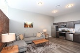 This thoughtfully designed single-floor unit blends contemporary style with functional living. The open-concept layout connects the kitchen, dining, and living areas, creating a cohesive and airy atmosphere throughout. The kitchen is outfitted with dark espresso cabinetry, granite countertops, and a full suite of stainless steel appliances, including a refrigerator, electric range, microwave, and dishwasher, all set above durable wood-look flooring. A dedicated dining area accommodates a round table and chairs, complemented by a striking floor-to-ceiling textured wood accent wall that adds warmth and visual interest to the open living space. The living room features a generously sized sectional sofa, a live-edge wood coffee table, and a glass door providing access to an outdoor area, along with clerestory windows that draw in natural light. The bedroom is a serene retreat, highlighted by the same rich wood accent wall serving as a dramatic headboard backdrop, plush carpeting, and a closet with built-in shelving. The bathroom is finished with granite countertops, dark wood cabinetry, a large vanity mirror, and a glass-enclosed walk-in shower, all unified by the same wood-look flooring found throughout the main living spaces.