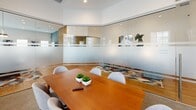 Located at 157 Hathaway Lane in Mooresville, NC, this polished conference room offers a refined and functional environment designed for professional gatherings. The single-room space is defined by its floor-to-ceiling frosted and clear glass partition walls, which provide both privacy and an open, transparent aesthetic that connects the room visually to the surrounding area. A warm-toned rectangular wood conference table serves as the room's centerpiece, surrounded by comfortable upholstered gray chairs that accommodate a small group. Recessed lighting in the white ceiling complements the abundant natural light that filters through multiple large windows fitted with white horizontal blinds, offering views of the exterior building surroundings. The flooring transitions between neutral carpet and hardwood, adding textural contrast to the clean, bright interior. A wall-mounted flat-screen display is positioned for easy viewing during presentations. Contemporary artwork adorns the white walls, lending color and personality to the otherwise understated palette. The overall ambiance is crisp, modern, and professional — a well-appointed space that balances functionality with thoughtful design details, making it well-suited for meetings, presentations, or collaborative work sessions.