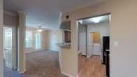 The Palm is a thoughtfully laid-out 2-bedroom, 2-bathroom single-story residence that balances functionality with comfortable living. The open-concept living area features plush carpeting, crown molding, and a ceiling fan, with natural light streaming in through windows and a glass door that leads to an outdoor space framed by greenery. The kitchen is designed for efficiency, featuring warm oak cabinetry, laminate countertops, hardwood-style flooring, and a full suite of stainless steel appliances including a refrigerator, range, and over-the-range microwave. A peninsula connects the kitchen to the living area, creating a practical flow between spaces. A dedicated in-unit laundry room with a full-size washer and dryer adds everyday convenience. Both bedrooms are carpeted and include ceiling fans and windows with outdoor views. Each bathroom offers hardwood-style flooring, oak vanity cabinetry, and walk-in closet access with wire shelving systems for ample storage. One bathroom includes a tiled soaking tub, while the other features a vanity with overhead cabinetry and a large mirror. The overall aesthetic is warm and neutral throughout.