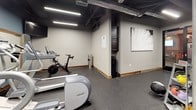 This fitness center offers a comprehensive workout environment designed to accommodate a variety of exercise routines. The space features durable speckled rubber flooring throughout, providing both safety and sound absorption during workouts. The exposed ceiling with visible ductwork and structural columns lends an industrial aesthetic, while linear LED lighting ensures the area is well-illuminated.

The cardio section is equipped with multiple Precor machines, including ellipticals and treadmills, each featuring individual display screens. A stationary spin bike rounds out the cardio offerings. Large wood-framed mirrors line the walls, enhancing the sense of depth and allowing users to monitor their form during exercise.

The free weights and functional training area includes medicine balls, kettlebells, a stability ball, and a dedicated storage rack. Dark cabinetry provides organized storage, and a water cooler offers convenient hydration. A wall-mounted television and a striking black-and-white architectural art piece add character to the space. A split-system air conditioning unit ensures comfortable temperature regulation throughout workouts. The overall ambiance is modern yet industrial, creating a focused and motivating environment.