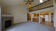 This two-story home makes an immediate impression with its dramatic vaulted ceilings that soar above the main living area, creating an expansive, airy atmosphere. A ceiling fan is centrally positioned overhead, while a skylight above the loft level draws in additional natural light. The living room is anchored by a stone fireplace, adding a warm and classic architectural element to the space. Soft carpeting throughout the main floor contributes to a comfortable and inviting ambiance.

The open-concept design connects the living area seamlessly to the kitchen, which features warm honey-toned wood cabinetry, recessed lighting, and a peninsula with a sink that doubles as a breakfast bar. The kitchen also includes white appliances and tile flooring, offering a clean and practical workspace.

A loft level overlooks the main living area from above, accessible by a staircase with a classic wood-railed balustrade. This upper level adds functional square footage while enhancing the grand, open feel of the home's interior. The two-floor layout balances openness on the main level with the privacy afforded by the elevated second floor, making this a well-designed space suited for comfortable everyday living.