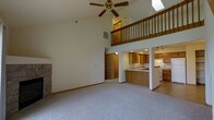 This two-story home makes an immediate impression with its dramatic vaulted ceilings that soar above the main living area, creating an expansive, airy atmosphere. A ceiling fan is centrally positioned overhead, while a skylight above the loft level draws in additional natural light. The living room is anchored by a stone fireplace, adding a warm and classic architectural element to the space. Soft carpeting throughout the main floor contributes to a comfortable and inviting ambiance.

The open-concept design connects the living area seamlessly to the kitchen, which features warm honey-toned wood cabinetry, recessed lighting, and a peninsula with a sink that doubles as a breakfast bar. The kitchen also includes white appliances and tile flooring, offering a clean and practical workspace.

A loft level overlooks the main living area from above, accessible by a staircase with a classic wood-railed balustrade. This upper level adds functional square footage while enhancing the grand, open feel of the home's interior. The two-floor layout balances openness on the main level with the privacy afforded by the elevated second floor, making this a well-designed space suited for comfortable everyday living.