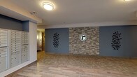The lobby at Meridia Transit Crossing presents a thoughtfully designed communal entrance space that balances contemporary architecture with warm, welcoming details. Wood-look tile flooring runs throughout, lending a cohesive and clean aesthetic across the entire area.

The main entrance features floor-to-ceiling black-framed glass doors and expansive windows that flood the space with natural light, offering views of the surrounding exterior. A seating area nearby includes a classic upholstered armchair, a tufted bench, and tall tropical houseplants, creating an inviting resting spot beneath soaring ceilings.

An accent wall finished in textured stacked stone, set against a muted blue-grey paint, adds visual depth and a striking focal point. Decorative wall art, including a botanical metal piece and a sculptural medallion arrangement, contributes to the refined character of the space.

A stainless steel elevator provides vertical access, positioned adjacent to a resident mailbox bank for added convenience. Flush-mount ceiling fixtures provide consistent ambient lighting throughout. The overall ambiance is modern yet approachable, with carefully curated furnishings and architectural details that establish a polished first impression for residents and visitors alike.