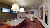 This dedicated game room offers a well-appointed space designed for leisure and social interaction. At its centerpiece sits a full-size billiards table, bathed in warm light from a pair of large, elegant pendant lamps that hang prominently overhead, ensuring excellent visibility during play. The room's bold, patterned carpet in deep reds and burgundy adds a dynamic visual energy underfoot, complementing the overall atmosphere.

A wall-mounted flat-screen television is positioned on one end of the room, making it easy to catch a game or broadcast while enjoying the space. On the opposite wall, a curated gallery of vibrant, colorful artwork lends an artistic and lively character to the environment.

Comfortable seating is thoughtfully arranged throughout the room, including upholstered armchairs and a chaise lounge, providing inviting spots for guests to relax and socialize between games. Recessed lighting supplements the pendant fixtures, keeping the space bright and welcoming. The neutral gray walls serve as a clean backdrop that allows the room's colorful accents — from the carpet to the artwork — to stand out. Overall, this game room strikes a balance between energetic recreation and relaxed comfort, making it a versatile gathering space.