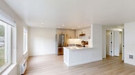 This thoughtfully designed 2-bedroom, 1-bathroom single-floor home offers a clean, modern aesthetic with a bright and cohesive interior. Light-toned hardwood flooring flows seamlessly throughout every room, creating a warm and unified feel. The open-concept living and kitchen area serves as the heart of the home, where a sleek kitchen island with a white countertop anchors the space. The kitchen is well-appointed with light gray flat-front cabinetry, black hardware accents, stainless steel appliances including a full-size refrigerator and range, under-cabinet lighting, and ample storage including open shelving. Recessed lighting illuminates the kitchen and living area, while a large front-facing window fills the living room with natural light and offers a view of the surrounding neighborhood. The primary bedroom is a comfortably sized retreat featuring a sliding-door closet with built-in hanging storage. The secondary bedroom is bright with a window and overhead light fixture. The full bathroom is finished in a minimalist style with white cabinetry, a clean countertop, a bathtub with shower, and black fixture accents that complement the overall design. Throughout the home, crisp white walls and streamlined finishes contribute to a fresh, contemporary ambiance.
