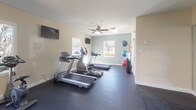 This fitness center offers a comprehensive workout environment designed to accommodate a wide range of exercise routines. The space features durable dark rubber flooring throughout, providing a safe and functional surface for all activities. Natural light streams in through multiple large windows that frame views of mature trees and landscaping, creating an inviting and energizing atmosphere. Ceiling fans help maintain comfortable airflow during workouts.

The cardio section is well-appointed with multiple treadmills and elliptical machines, complemented by wall-mounted flat-screen televisions for entertainment. The strength training area includes a multi-station cable machine, a rowing machine, and various resistance equipment to support a full-body workout. A dedicated free weights section offers a tiered kettlebell rack with a range of weights, while stability balls are neatly stored along the wall for functional training.

A large wall-to-wall mirror visually expands the space and aids in proper form during exercise. An open stretching and mat area features several floor exercise mats laid out for yoga, Pilates, or cool-down routines. The overall design balances functionality and comfort, making this fitness center a versatile and well-rounded facility for residents or members seeking a complete exercise experience.