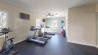 This fitness center offers a comprehensive workout environment designed to accommodate a wide range of exercise routines. The space features durable dark rubber flooring throughout, providing a safe and functional surface for all activities. Natural light streams in through multiple large windows that frame views of mature trees and landscaping, creating an inviting and energizing atmosphere. Ceiling fans help maintain comfortable airflow during workouts.

The cardio section is well-appointed with multiple treadmills and elliptical machines, complemented by wall-mounted flat-screen televisions for entertainment. The strength training area includes a multi-station cable machine, a rowing machine, and various resistance equipment to support a full-body workout. A dedicated free weights section offers a tiered kettlebell rack with a range of weights, while stability balls are neatly stored along the wall for functional training.

A large wall-to-wall mirror visually expands the space and aids in proper form during exercise. An open stretching and mat area features several floor exercise mats laid out for yoga, Pilates, or cool-down routines. The overall design balances functionality and comfort, making this fitness center a versatile and well-rounded facility for residents or members seeking a complete exercise experience.