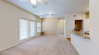 This single-floor residence offers a thoughtfully arranged layout with a comfortable, functional design throughout. The living area is generously sized, featuring soft neutral carpet, warm beige walls, and vaulted ceilings equipped with both a ceiling fan and track lighting. Large windows with horizontal blinds and a set of French doors allow abundant natural light to fill the space while providing outdoor access. A pass-through counter connects the living area to the kitchen, enhancing the open feel of the floor plan. The kitchen is well-appointed with warm honey-toned wood cabinetry, granite-style countertops, and white appliances including a refrigerator, electric range, and over-the-range microwave. Light hardwood flooring runs through the kitchen and connects seamlessly to adjacent spaces. A spacious walk-in closet is conveniently located just off the kitchen area, providing ample storage. The bedroom is carpeted and bright, featuring a ceiling fan and a pair of windows that invite natural light and outdoor views. The bathroom showcases hardwood flooring, warm wood vanity cabinetry, a white countertop with integrated sink, a large mirror, and a glass-enclosed shower, creating a clean and inviting atmosphere throughout.