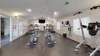 Located at 60 Knoll Ct in Carmel, Indiana, this well-equipped fitness center offers a comprehensive workout environment within a single, open-plan room. The space features vaulted ceilings that lend an airy, open feel, complemented by recessed lighting and a ceiling fan that ensures comfortable air circulation during workouts. Durable rubber flooring covers the entire area, providing both safety and practicality underfoot. Natural light filters in through multiple windows fitted with blinds, brightening the space throughout the day. A wall-mounted flat-screen television is positioned for easy viewing from cardio equipment. The room is outfitted with an array of cardio machines, including treadmills, ellipticals, and stationary bikes, as well as a dumbbell rack, weight bench, and resistance training equipment. A large wall mirror enhances the sense of space and allows for proper form checks during exercise. Resistance bands and exercise accessories are neatly organized on wall-mounted hooks and shelves. French doors with glass panels provide an elegant entry point, connecting the fitness center to adjacent spaces. Sanitation supplies and a waste bin are conveniently available, reflecting a well-maintained facility. Overall, this fitness center delivers a functional and inviting environment suited for a full range of exercise routines.