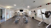 Located at 60 Knoll Ct in Carmel, Indiana, this well-equipped fitness center offers a comprehensive workout environment within a single, open-plan room. The space features vaulted ceilings that lend an airy, open feel, complemented by recessed lighting and a ceiling fan that ensures comfortable air circulation during workouts. Durable rubber flooring covers the entire area, providing both safety and practicality underfoot. Natural light filters in through multiple windows fitted with blinds, brightening the space throughout the day. A wall-mounted flat-screen television is positioned for easy viewing from cardio equipment. The room is outfitted with an array of cardio machines, including treadmills, ellipticals, and stationary bikes, as well as a dumbbell rack, weight bench, and resistance training equipment. A large wall mirror enhances the sense of space and allows for proper form checks during exercise. Resistance bands and exercise accessories are neatly organized on wall-mounted hooks and shelves. French doors with glass panels provide an elegant entry point, connecting the fitness center to adjacent spaces. Sanitation supplies and a waste bin are conveniently available, reflecting a well-maintained facility. Overall, this fitness center delivers a functional and inviting environment suited for a full range of exercise routines.