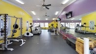 This fitness center is a comprehensive, single-floor facility designed to support a full range of workout routines. The interior is immediately distinguished by its bold color scheme — vibrant yellow and purple walls that create an energetic, motivating atmosphere throughout the space.

The facility is organized into distinct training zones. The cardio area features multiple elliptical machines and treadmills arranged in open rows, allowing ample space for movement. Ceiling fans and fluorescent lighting keep the environment comfortable and well-lit. Wall-mounted flat-screen televisions are positioned throughout, offering entertainment during workouts.

The strength training area is generously equipped with cable machines, weight benches, squat racks, and a full selection of plate-loaded equipment. A dedicated free weights section houses an extensive rack of dumbbells in varying sizes, accompanied by a weight bench for targeted exercises. The rubber flooring throughout provides durability and safety underfoot.

Large mirrors span entire walls across multiple zones, enhancing the sense of openness and allowing members to monitor their form. Additional functional training equipment, including stability balls, exercise mats, and plyometric boxes, is also available. The textured ceiling and multiple ceiling fans contribute to a practical, no-frills atmosphere focused entirely on fitness performance.