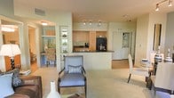 This single-floor space showcases an open-concept layout that connects the living, dining, and kitchen areas into one cohesive environment. The kitchen is equipped with light maple cabinetry, a dark countertop peninsula that doubles as a breakfast bar, a black refrigerator, and track lighting overhead, providing both functionality and a modern aesthetic. The peninsula creates a natural dividing point between the kitchen and the main living area while maintaining an open sightline throughout the space.

The living area features plush carpeting in a neutral tone, complemented by upholstered armchairs with decorative accent pillows, contributing to a comfortable and well-curated atmosphere. A partial wall with built-in display shelving adds architectural interest and serves as a subtle separator between the dining nook and main living area. The dining nook, visible to the left, is furnished with animal-print accent chairs, adding a touch of personality. Hardwood-style flooring is noted in transitional areas. A hallway visible toward the rear leads to additional rooms. The overall ambiance is light, airy, and well-organized.