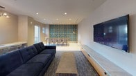 Located at 333 S Glebe Rd in Arlington, Virginia, this single-floor amenity space is thoughtfully designed with a cohesive contemporary aesthetic that blends bold color accents with refined materials. The welcoming lobby serves as a striking introduction, anchored by a sculptural gold multi-bulb chandelier suspended above a seating arrangement featuring a tufted gray sofa, teal accent chairs, and a marble-topped coffee table. A decorative geometric lattice screen wall and rich wood-paneled walls frame the space, while terrazzo flooring adds a polished, durable foundation.

Adjacent lounge and gathering areas continue the design language, incorporating patterned carpeting, a tufted navy sofa, large-format television, and built-in media cabinetry. A bold teal-and-gold geometric accent wall anchors the dining zone, which includes a communal table with seating for multiple guests.

The catering kitchen is outfitted with warm wood cabinetry, expansive marble-look quartz countertops, a white subway tile backsplash, and geometric gold pendant lighting. A separate conference or meeting room features floor-to-ceiling teal upholstered acoustic panels, a wood-slat ceiling, a central conference table, and a wall-mounted display screen—ideal for private gatherings or presentations. Natural light filters through windows in several areas, enhancing the overall bright and welcoming ambiance.
